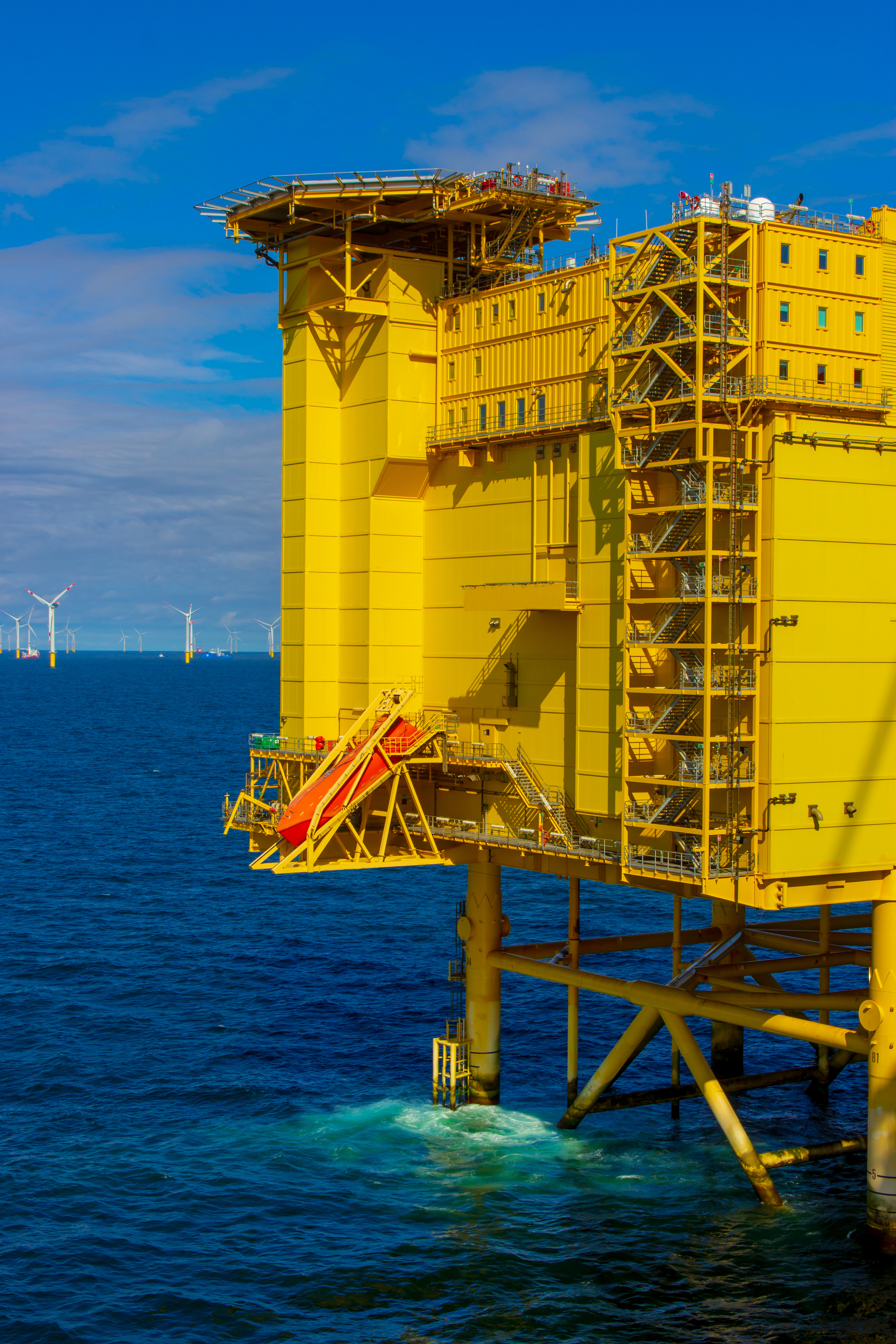 Teilansicht einer gelben Offshore-Wind-Gleichstrom-Plattform mit Windturbinen im Hintergrund.