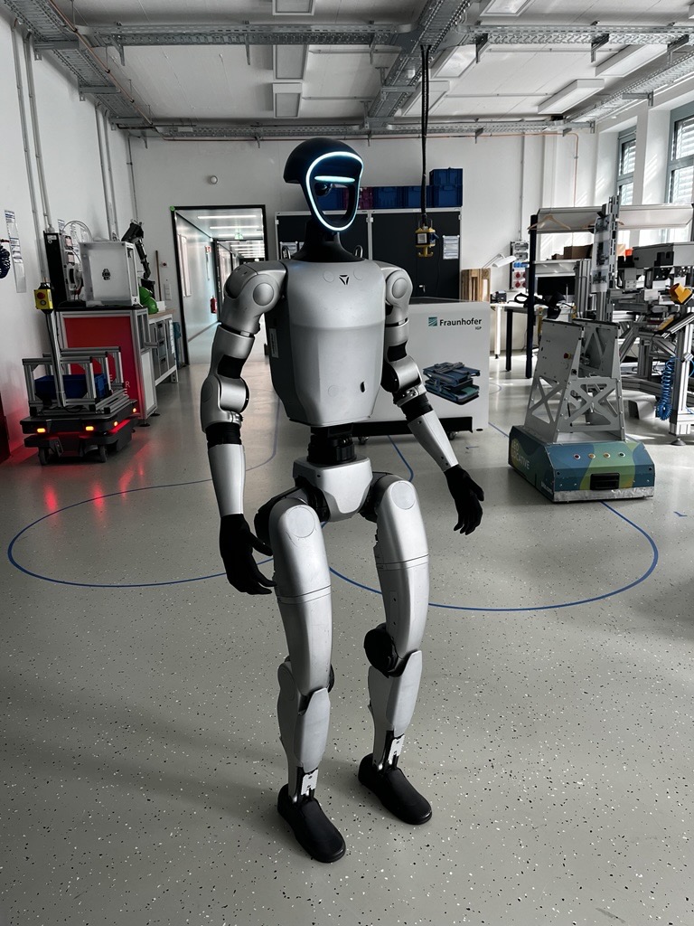 Image of a silver-coloured humanoid robot standing in a large room full of pieces of machinery at Fraunhofer IGP.