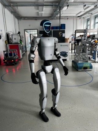 Image of a silver-coloured humanoid robot standing in a large room full of pieces of machinery at Fraunhofer IGP.